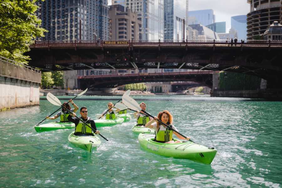 People in colorful kayaks paddling on the Chicago River between skyscrapers.