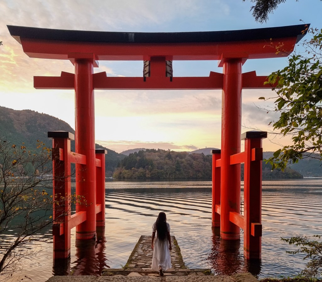 A large red torii gate standing in the water of a lake at sunrise with soft morning light.