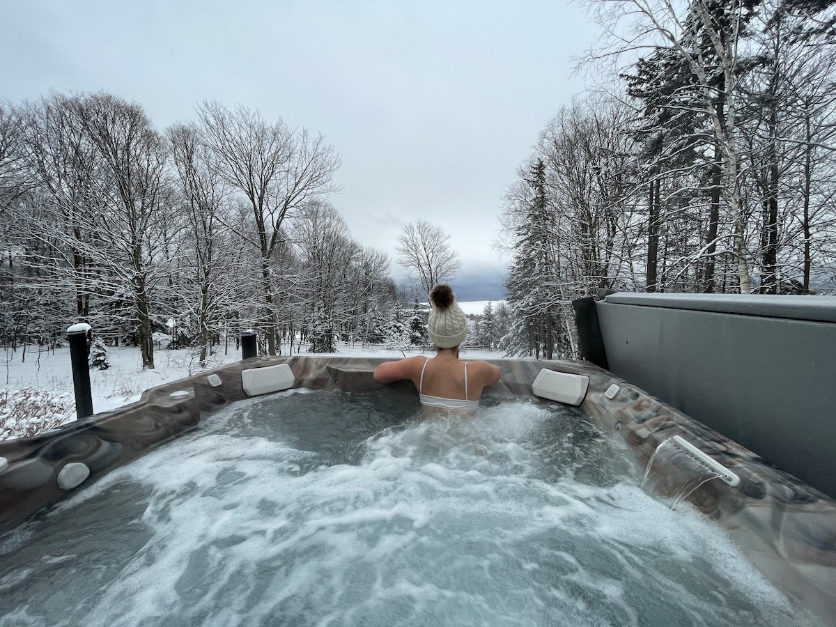 An outdoor steaming hot tub on a deck overlooking a snowy reservoir and forest.