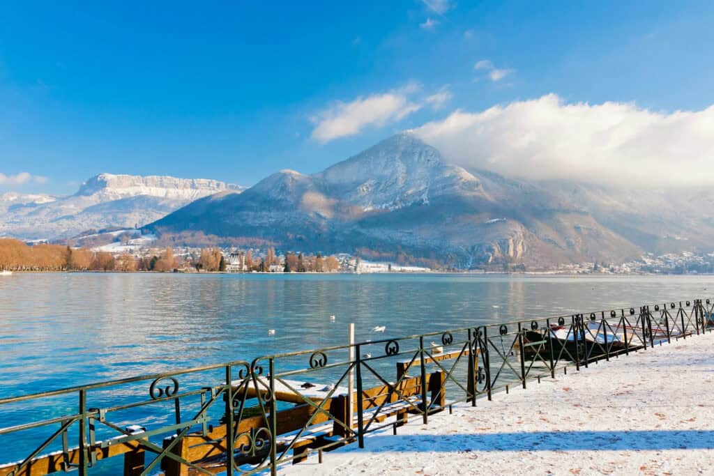 A wide winter panorama of Lake Annecy with clouds resting on the surrounding snow-topped mountain peaks.