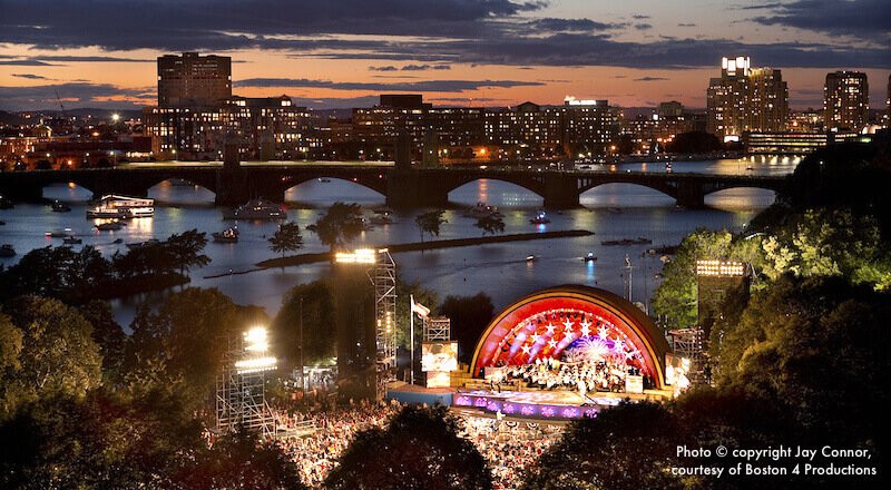 A large crowd gathered on the Charles River Esplanade facing the Hatch Shell stage during the day.