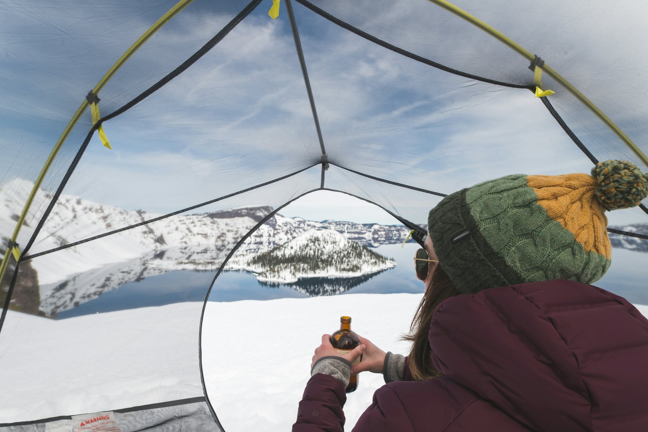 A hiker resting inside a tent pitched in the snow overlooking the partially frozen Crater Lake.