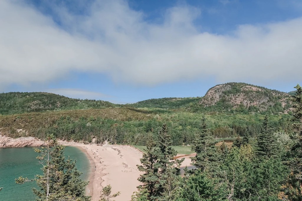 Aerial perspective of Sand Beach nestled between evergreen forests and rocky Acadia cliffs.