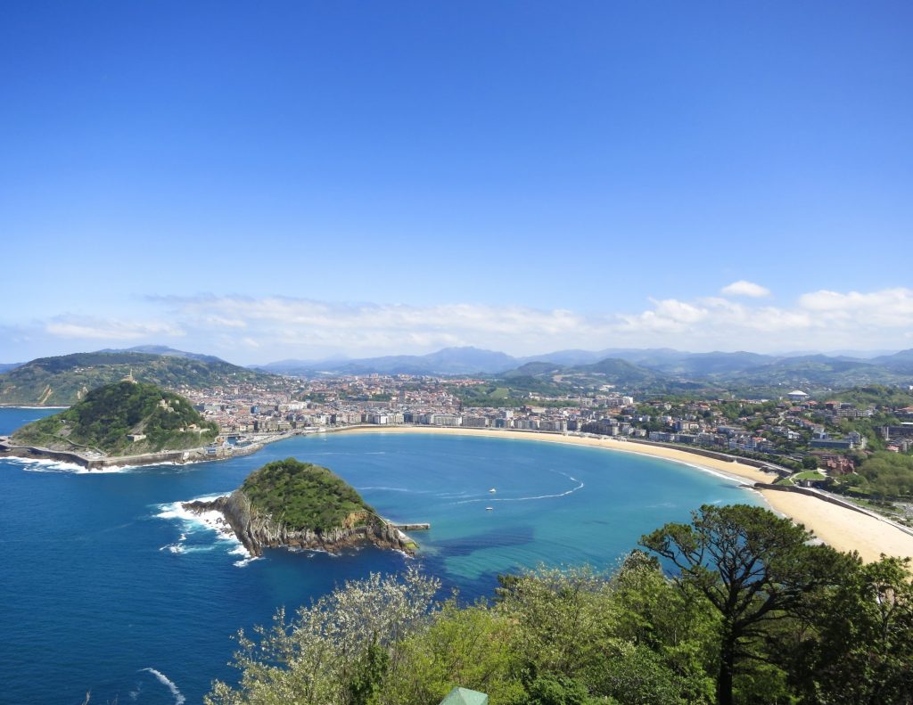 A wide view of a crescent-shaped beach and surrounding mountains under a soft sky.