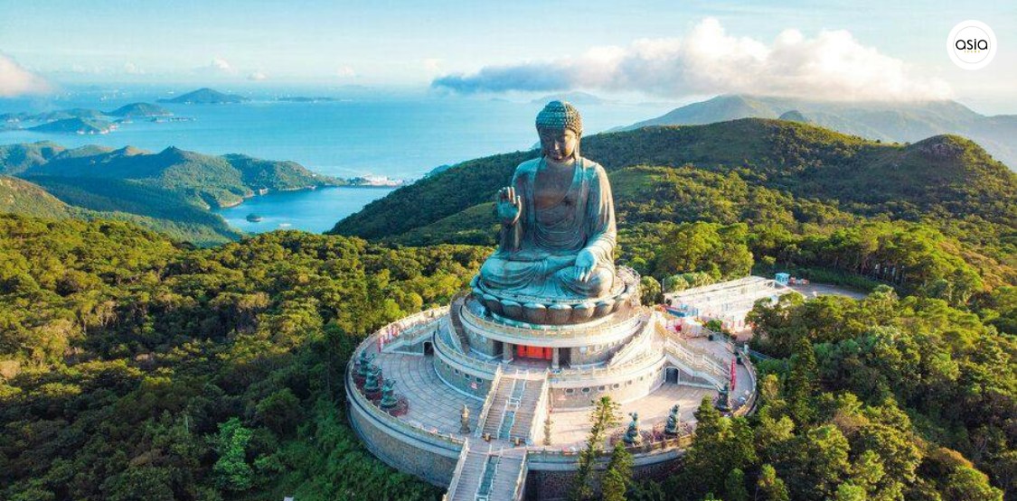Hikers walking along a narrow ridge trail overlooking a blue bay and green coastal mountains.