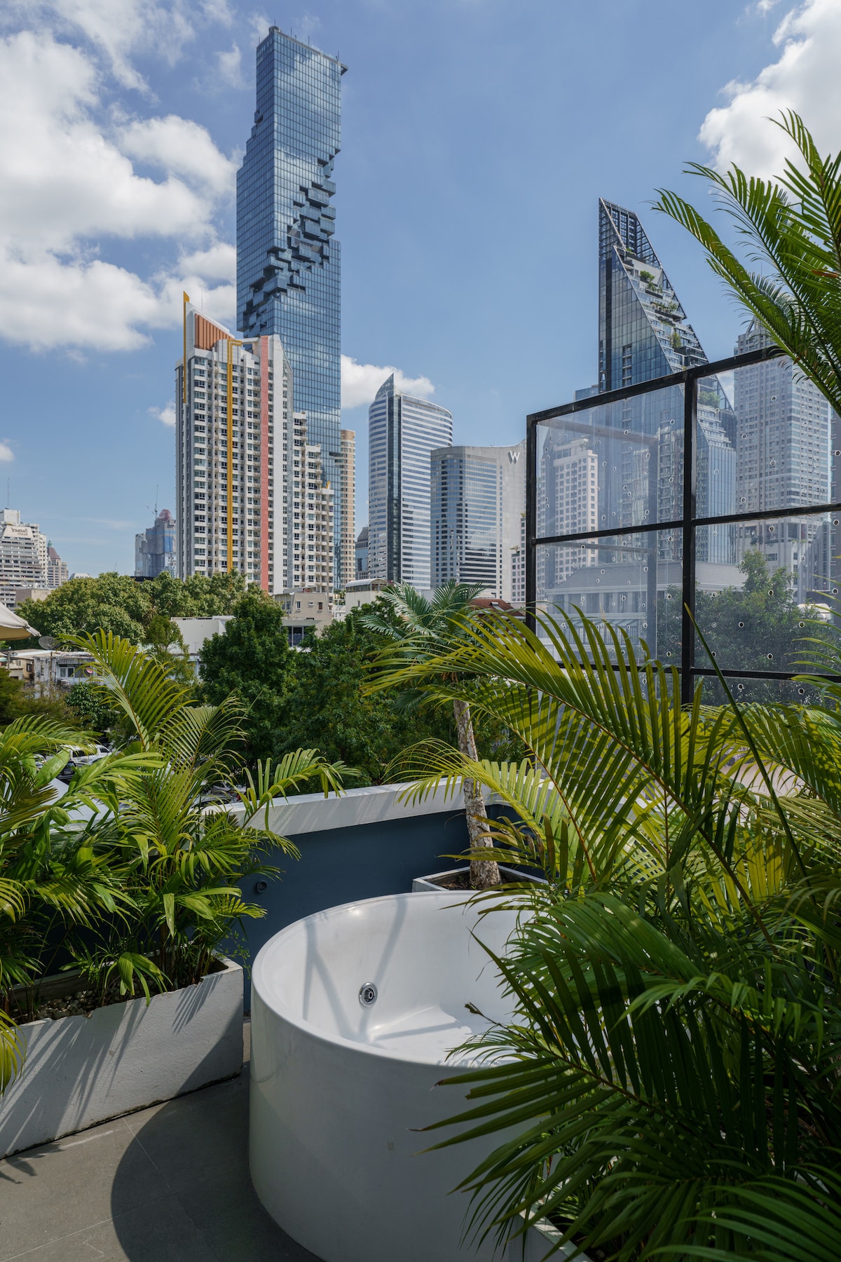 An outdoor rooftop jacuzzi surrounded by plants with the pixelated Mahanakohn building in the background.