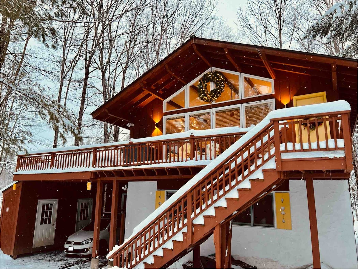 Exterior of a two-story wooden ski chalet with glowing windows at dusk and snow on the ground.