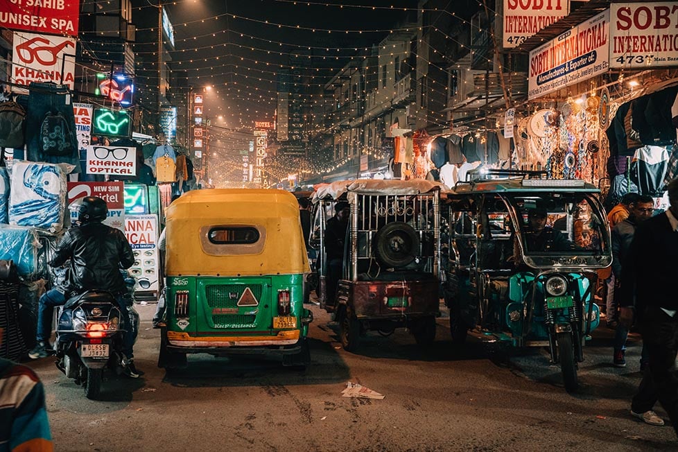 A yellow and green auto-rickshaw navigating through a crowded, busy street in Delhi.