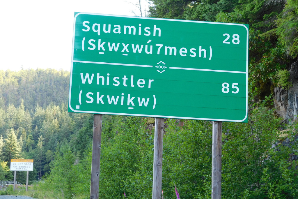 A green road sign on the Sea to Sky Highway showing place names in both English and Sḵwx̱wú7mesh.
