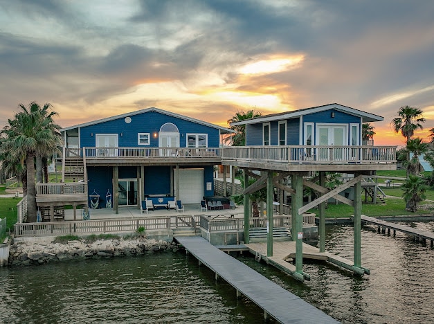 A blue waterfront vacation home with a private fishing pier during a vibrant sunset.
