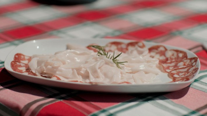 A rustic platter of sliced lardo and Tuscan salami on a red-checkered cloth.
