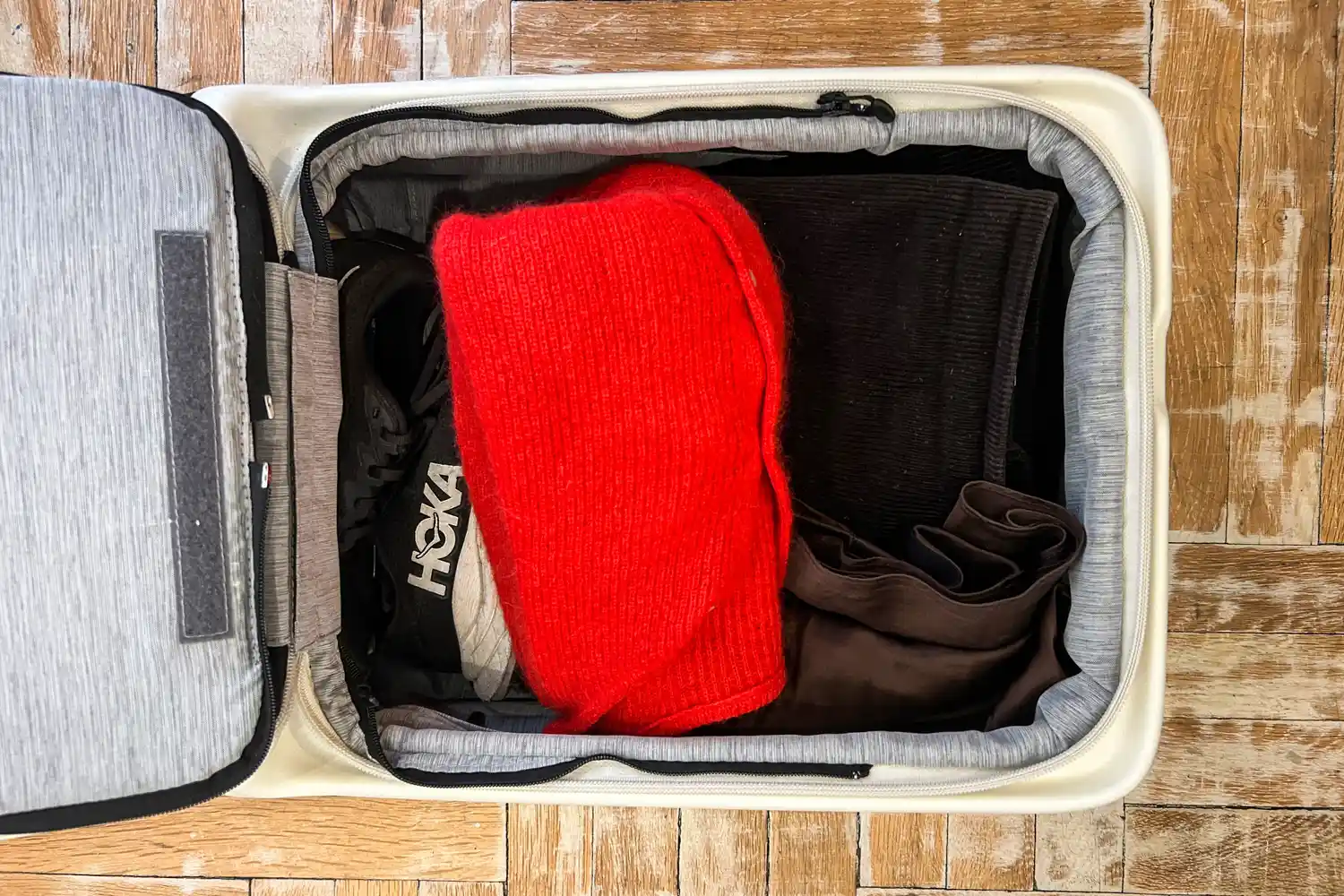 A carry-on suitcase partially packed with folded clothes and shoes on a wooden floor.