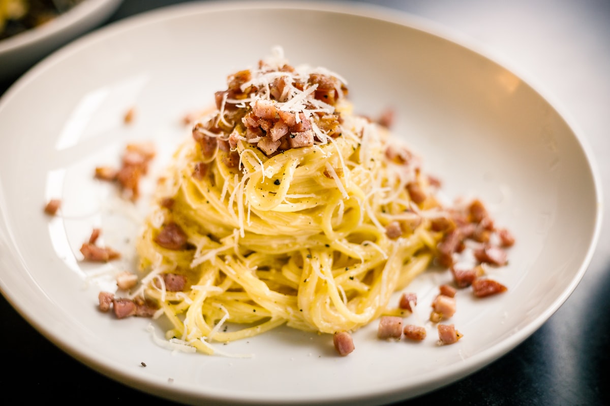 A close-up of a plate of creamy Rigatoni Carbonara garnished with black pepper and pecorino cheese.