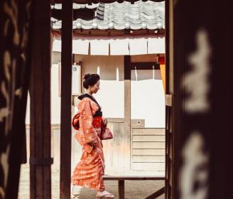 A woman in traditional Geisha attire walking through a historic Japanese setting.