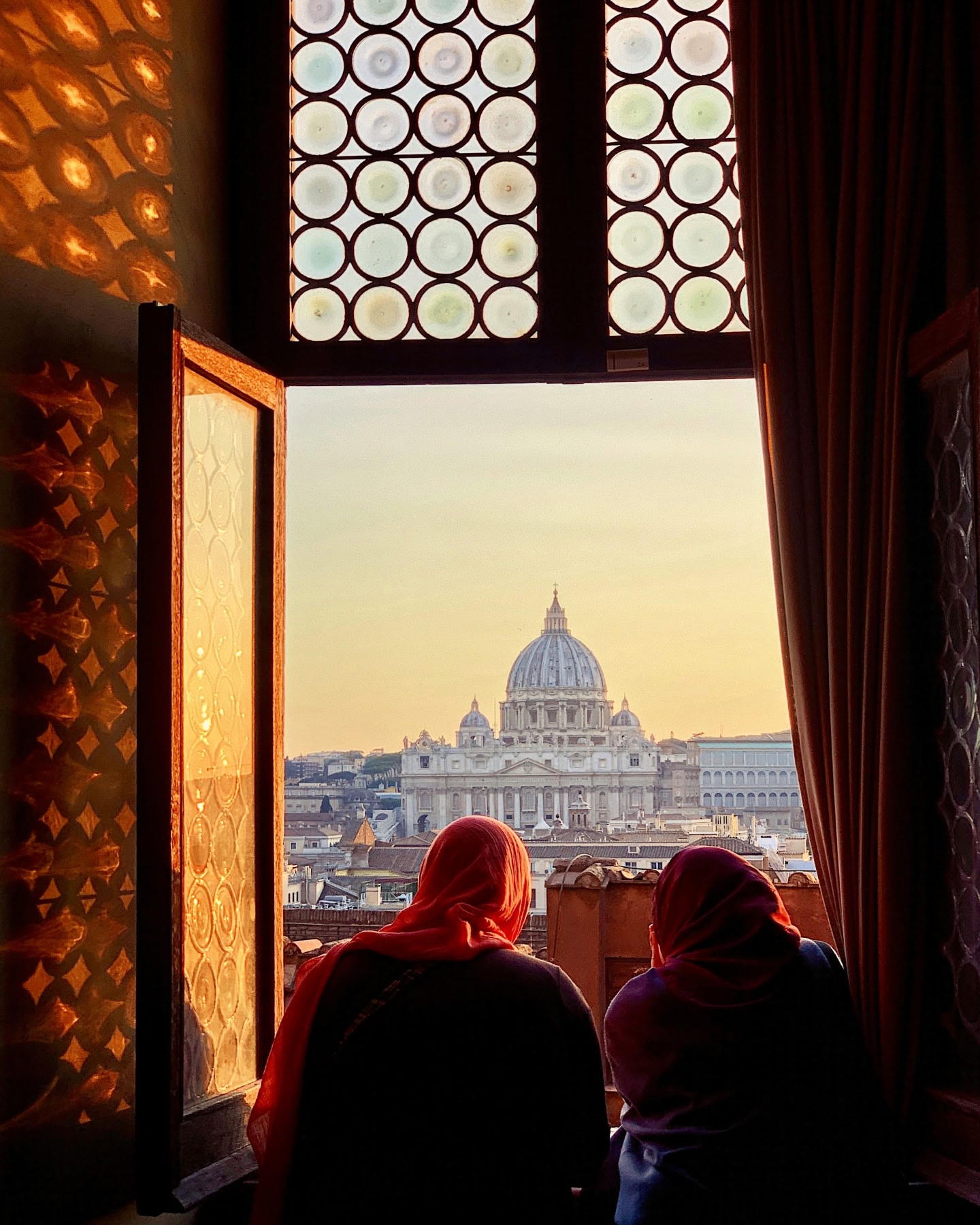 Two women standing on a terrace overlooking the St. Peter's Basilica dome in Rome during golden hour.