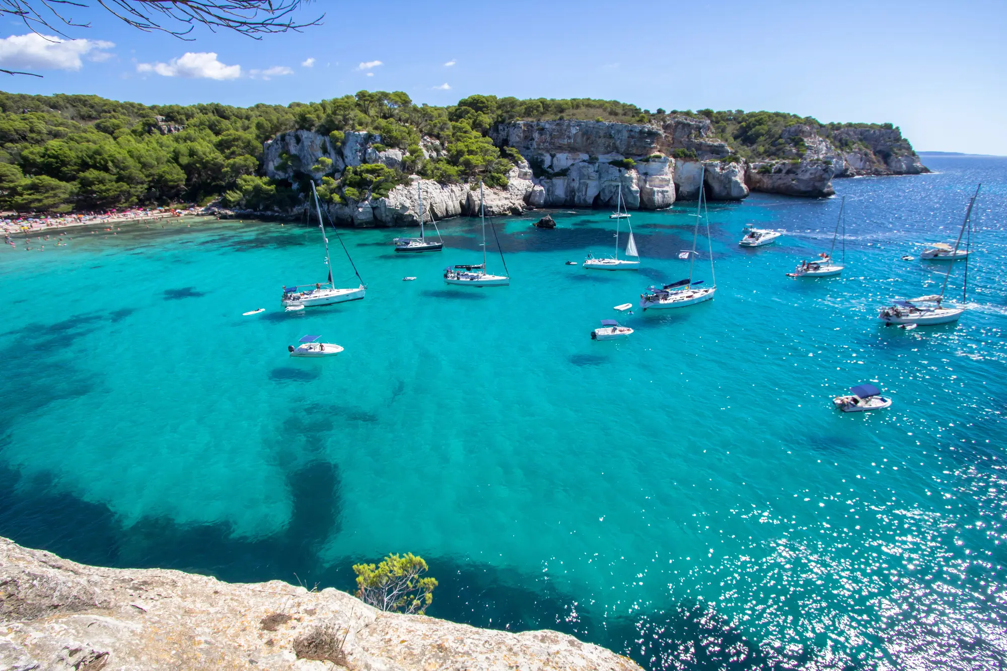 Several small leisure boats floating on extremely transparent turquoise water in a rocky Menorcan cove.