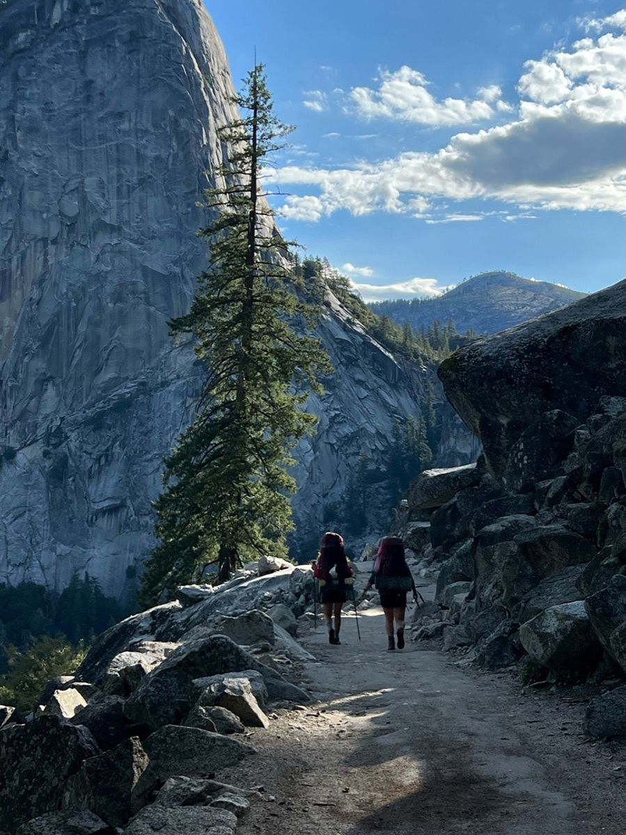 Two hikers traversing a scenic trail with Yosemite's granite peaks in the background.