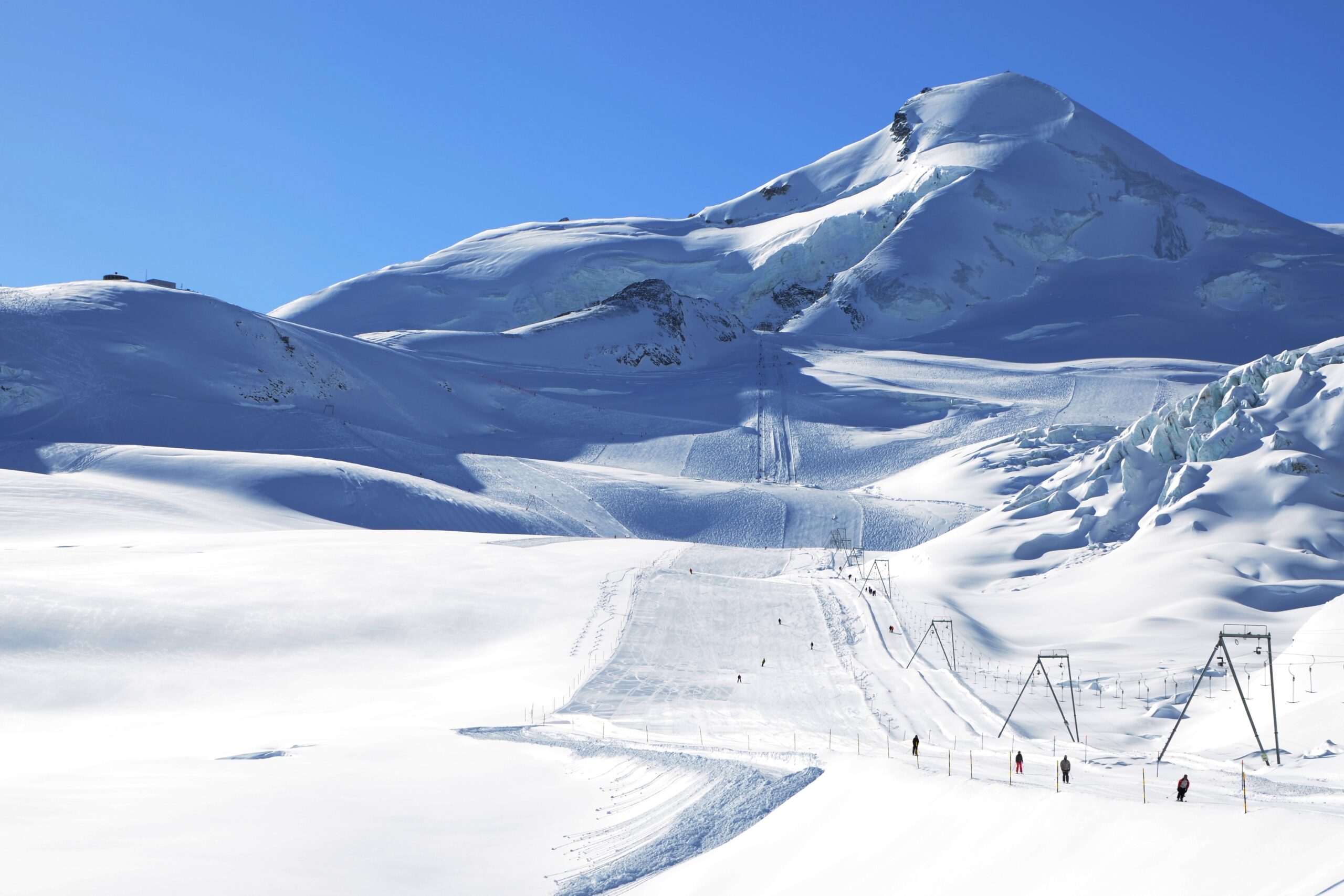 Skiers on a wide, high-altitude groomed slope with massive mountain peaks behind them.