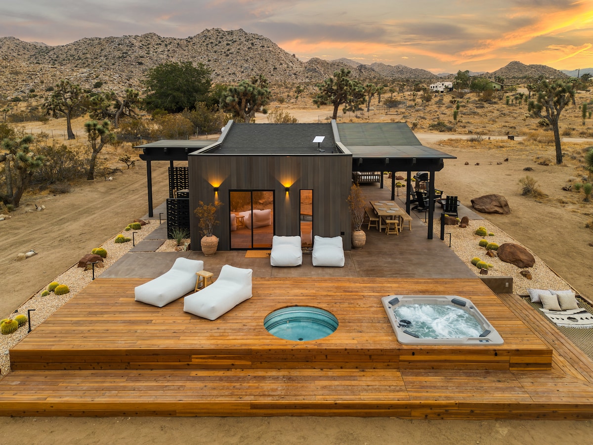 A modern desert home at dusk with warm interior lights and a glowing hot tub on the deck.