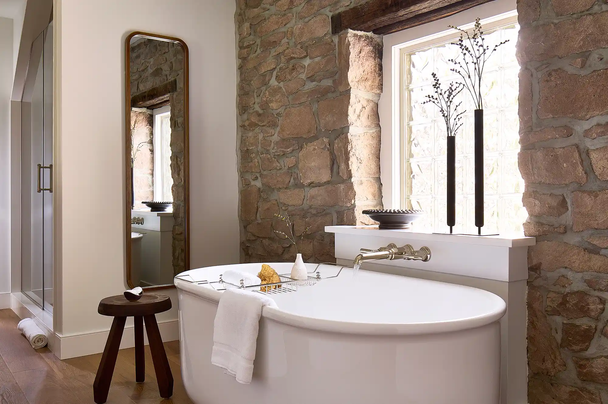 A modern freestanding bathtub situated against an original stone wall under a window.