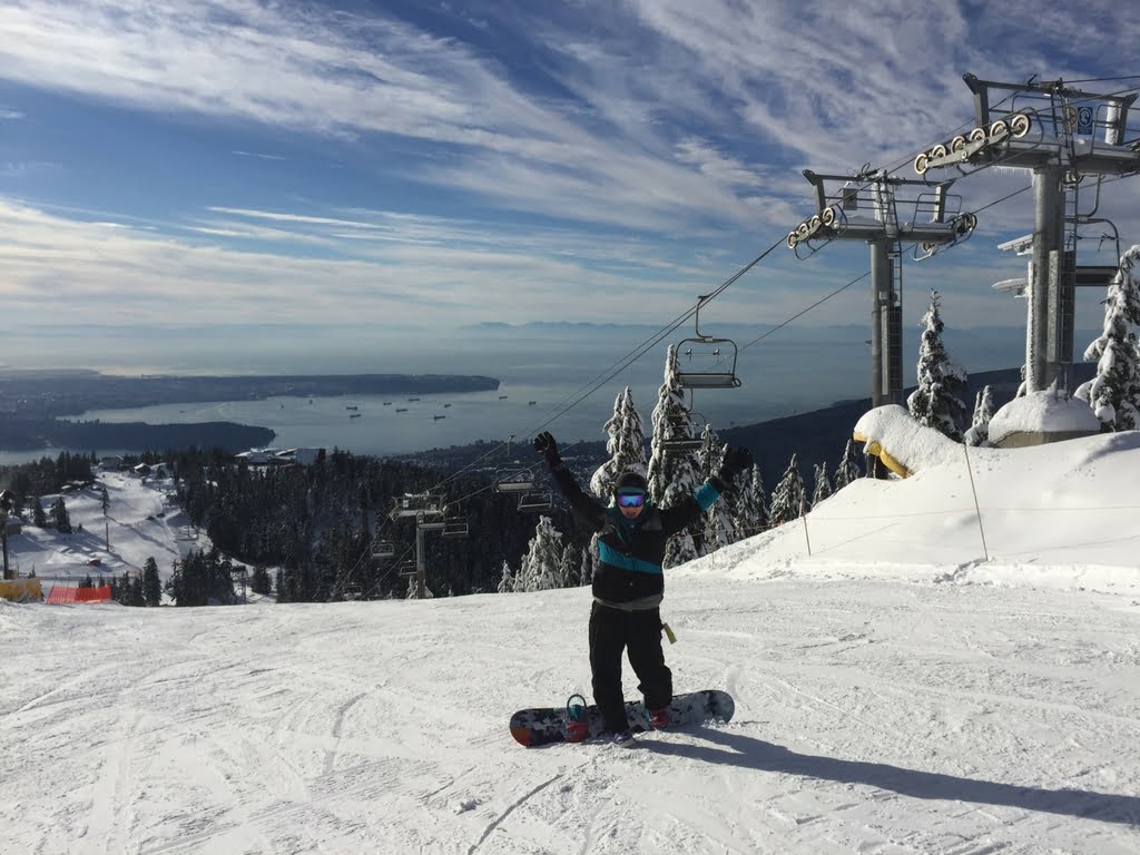 A snowboarder posing at the summit of Grouse Mountain with the city and ocean in the distance.