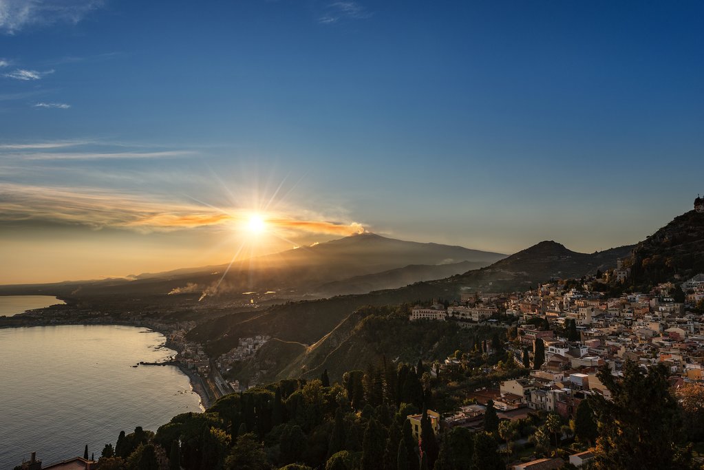 A panoramic view of a Sicilian coastal town with historic buildings and deep blue Mediterranean waters.