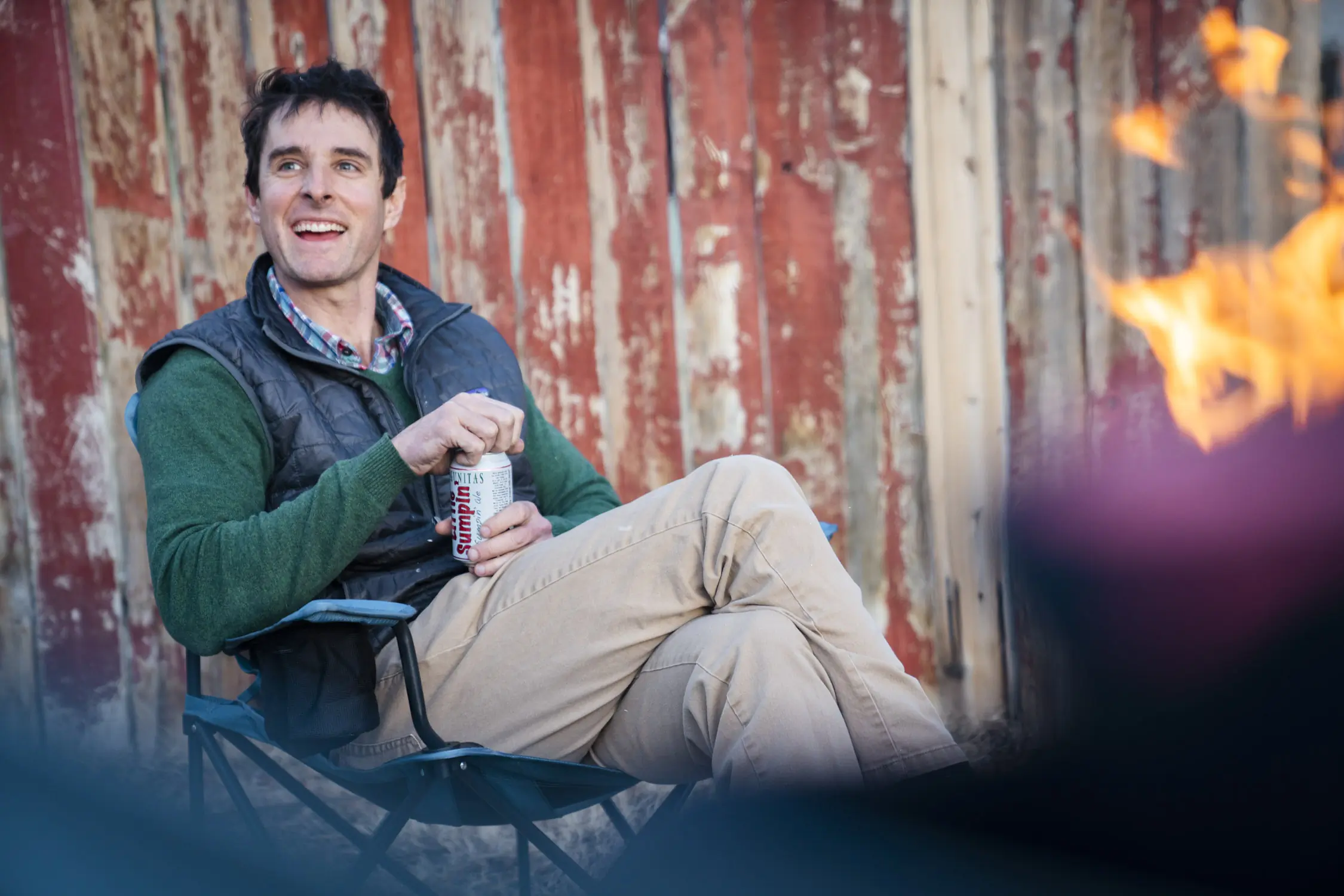 A man relaxing in a blue REI Co-op camping chair next to a small stone campfire.