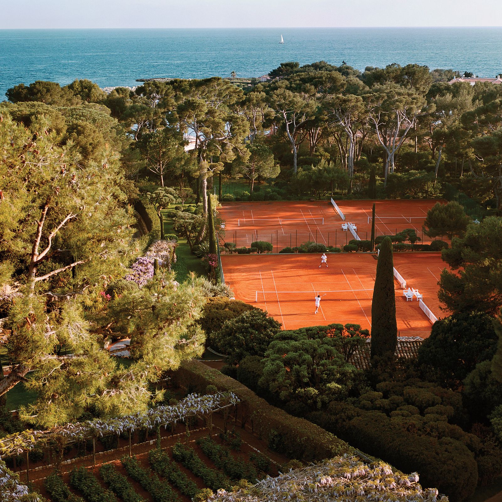 A luxury tennis court surrounded by lush greenery and ocean views at a high-end French hotel.