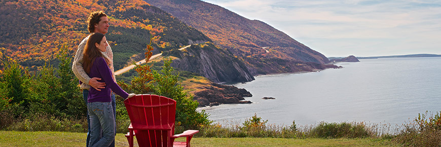 Two people sitting in red chairs on a cliffside overlooking the Cabot Trail and the ocean.