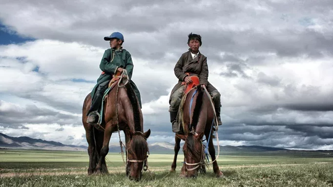 Two young Mongolian boys riding horses across the grassy plains.