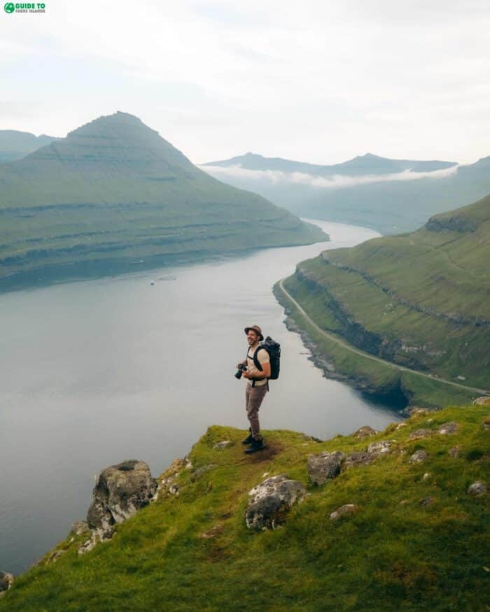 A person sitting on a mountain ridge overlooking a deep blue fjord and steep green peaks.