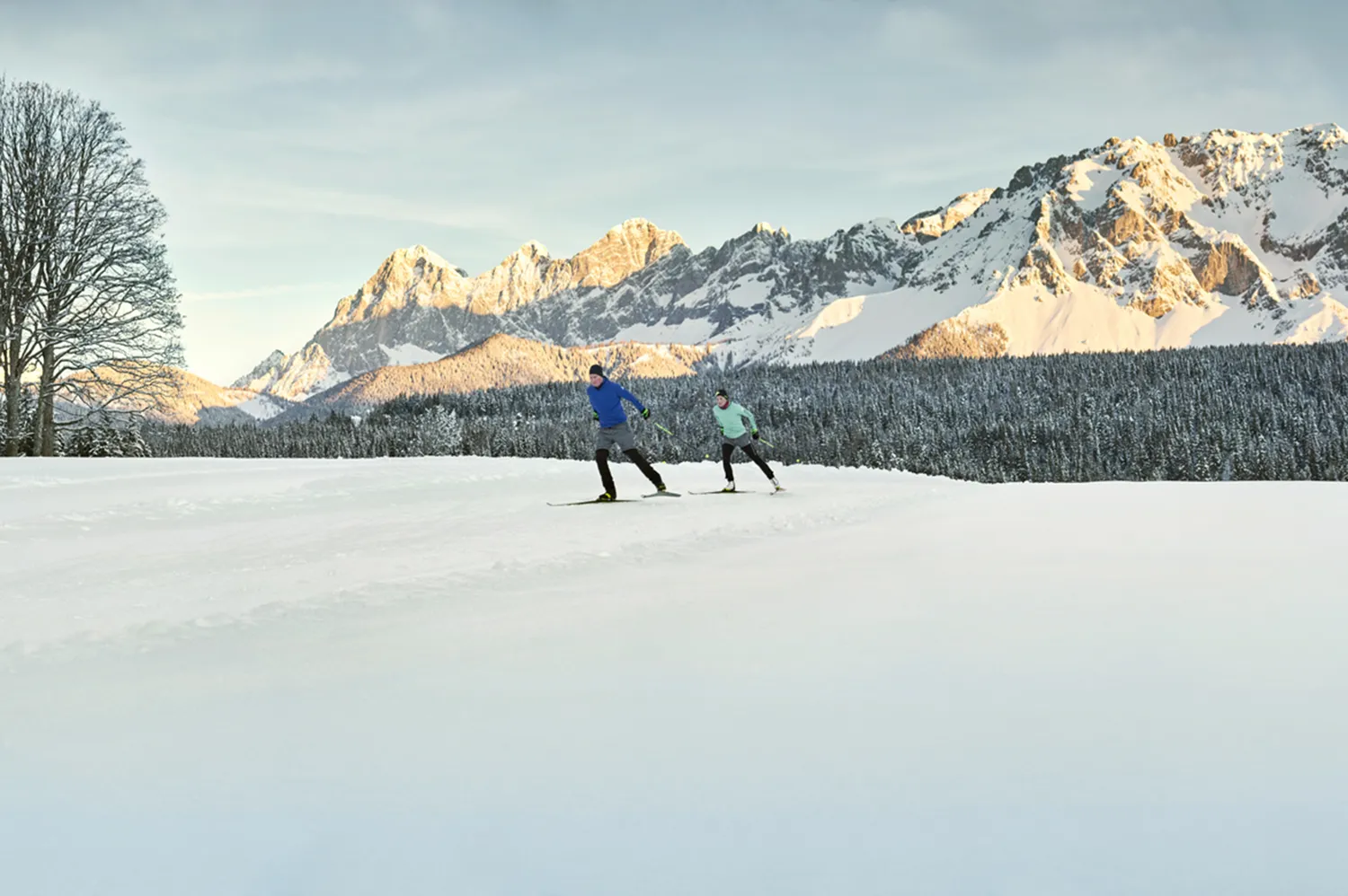 Two people cross-country skiing across a vast, flat snowy plateau with dramatic mountain ranges behind them.