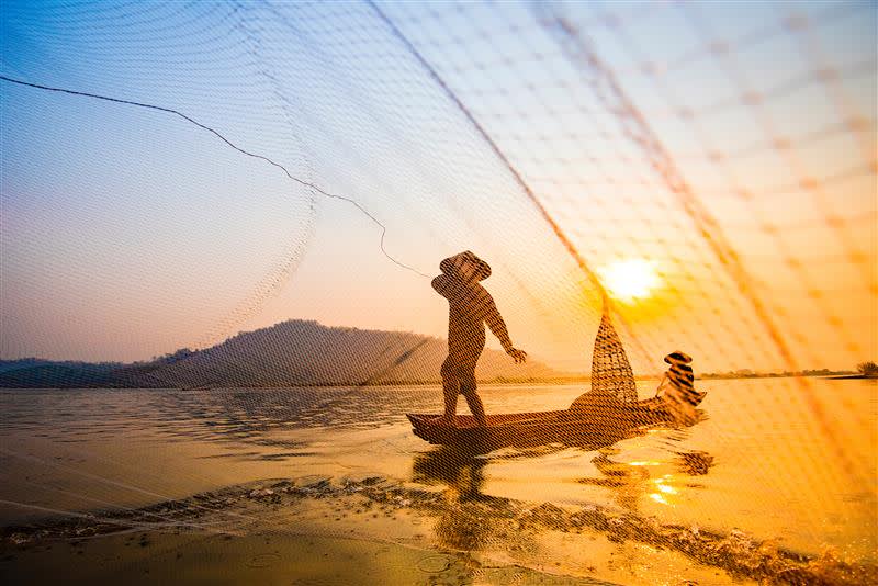 A traditional fisherman casting a circular net over the Mekong River at sunset.