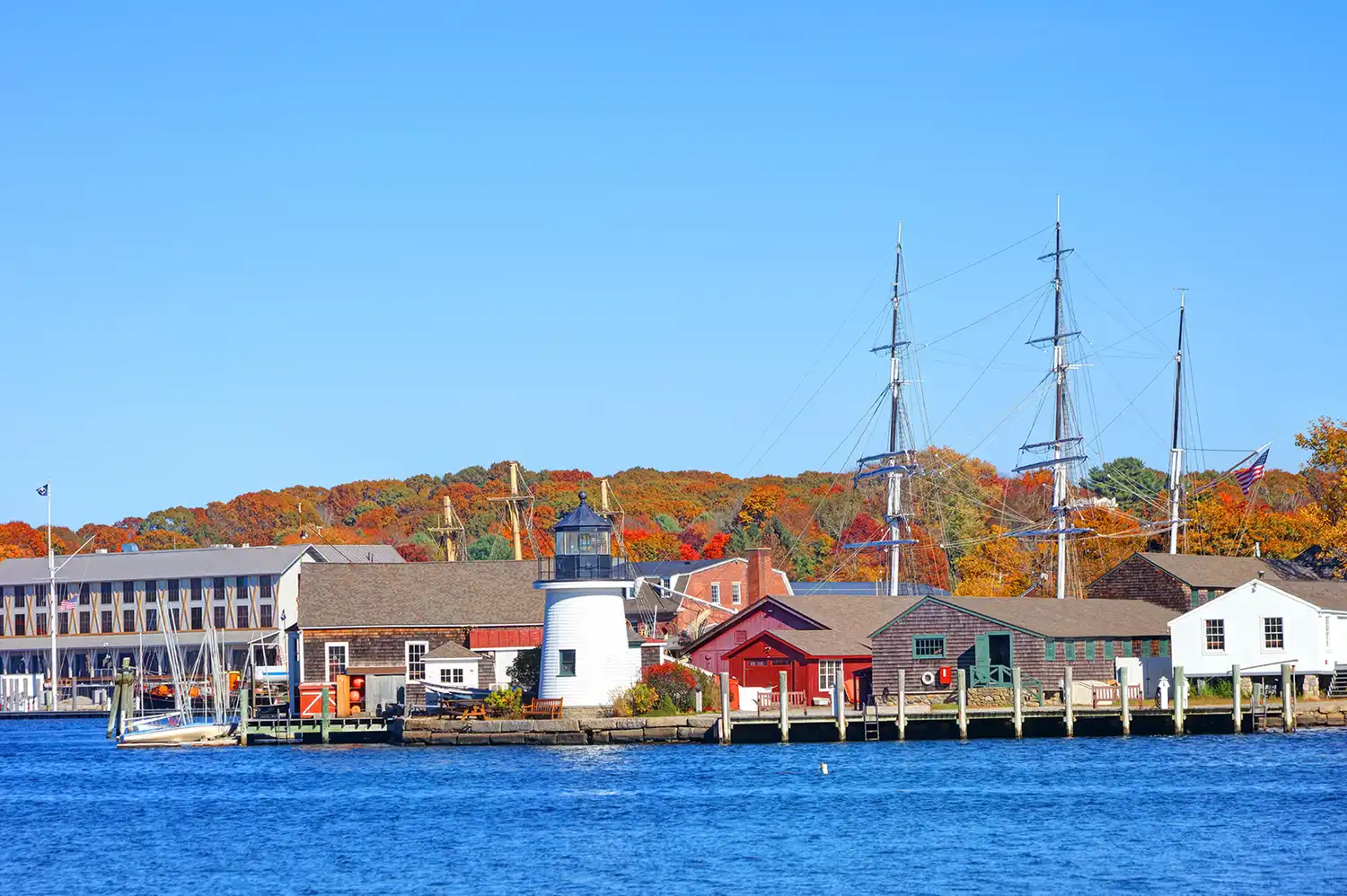 The Mystic harborfront featuring a historic lighthouse, a sailing ship, and autumn trees.