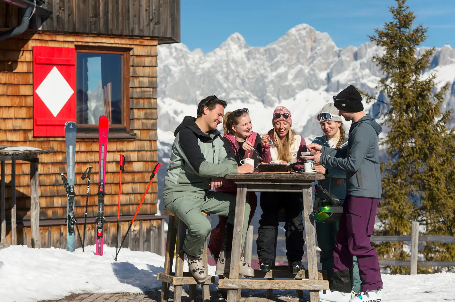 A group of skiers sitting at a rustic wooden table outside a mountain hut with snowy peaks in the background.