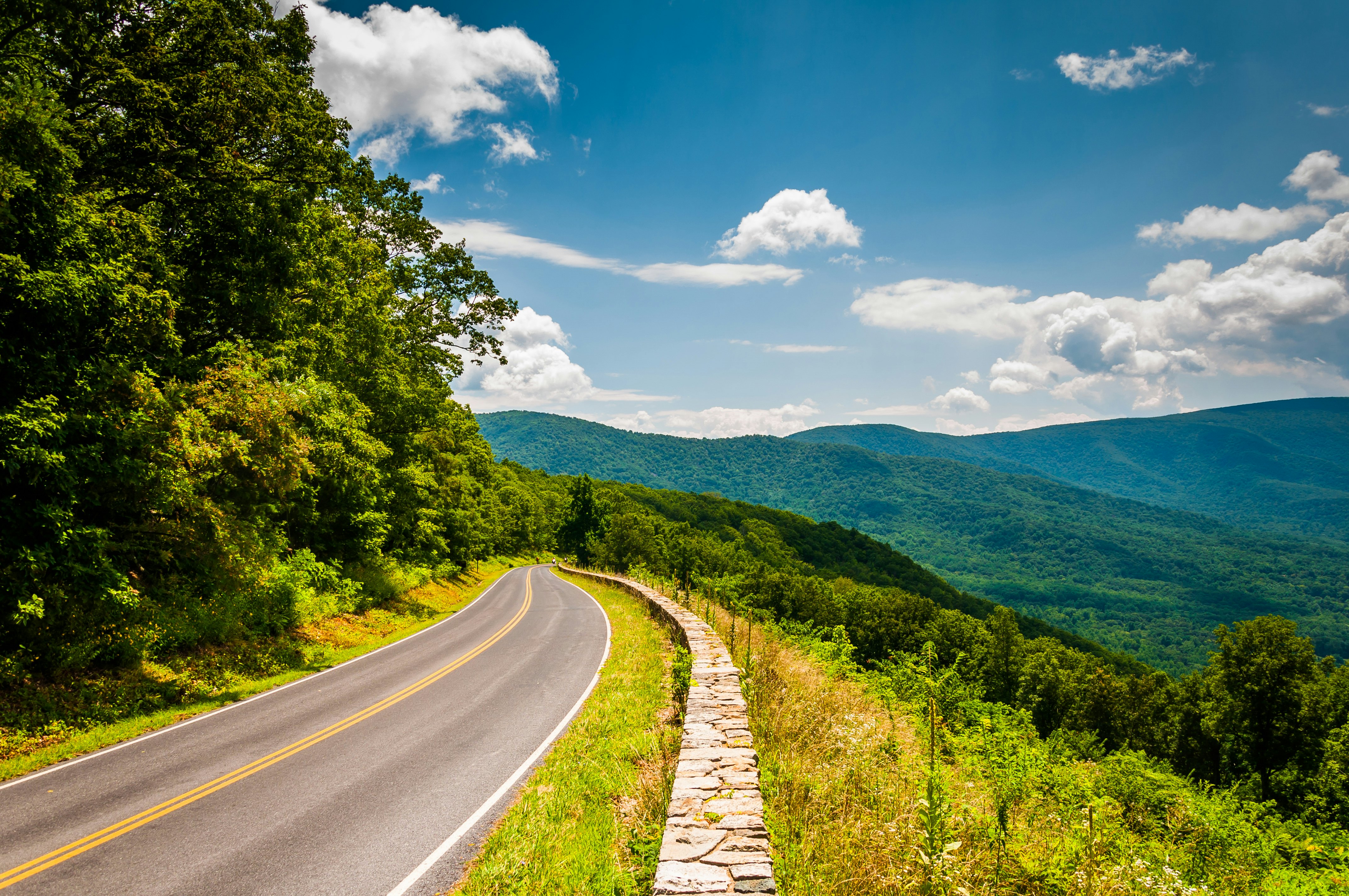 A two-lane mountain road with a historic stone wall overlooking distant rolling blue ridges.