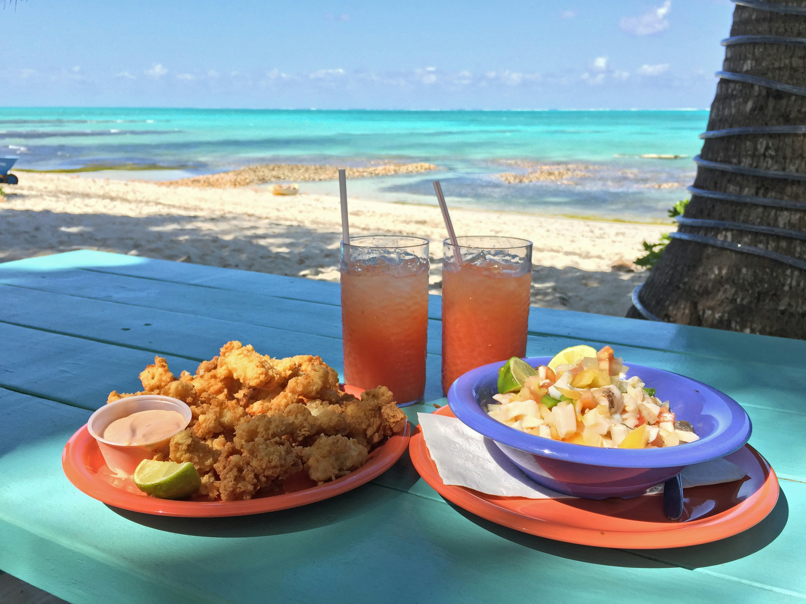 Plates of fresh Bahamian conch salad and cracked conch served on a blue table overlooking the ocean.