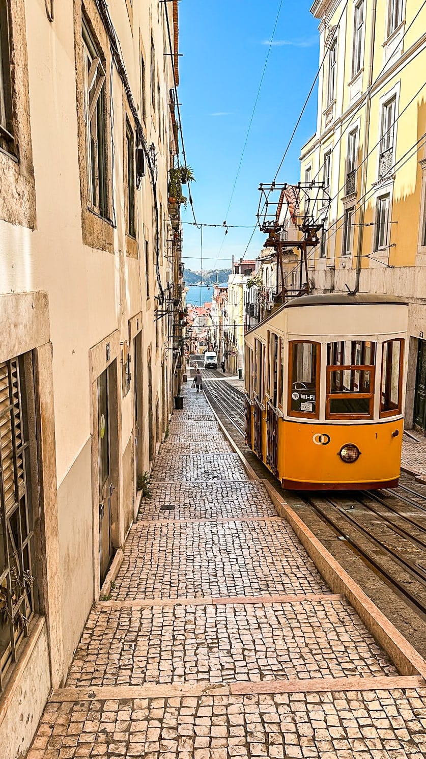 A traditional yellow tram climbing a steep cobblestone hill in a historic Lisbon neighborhood.