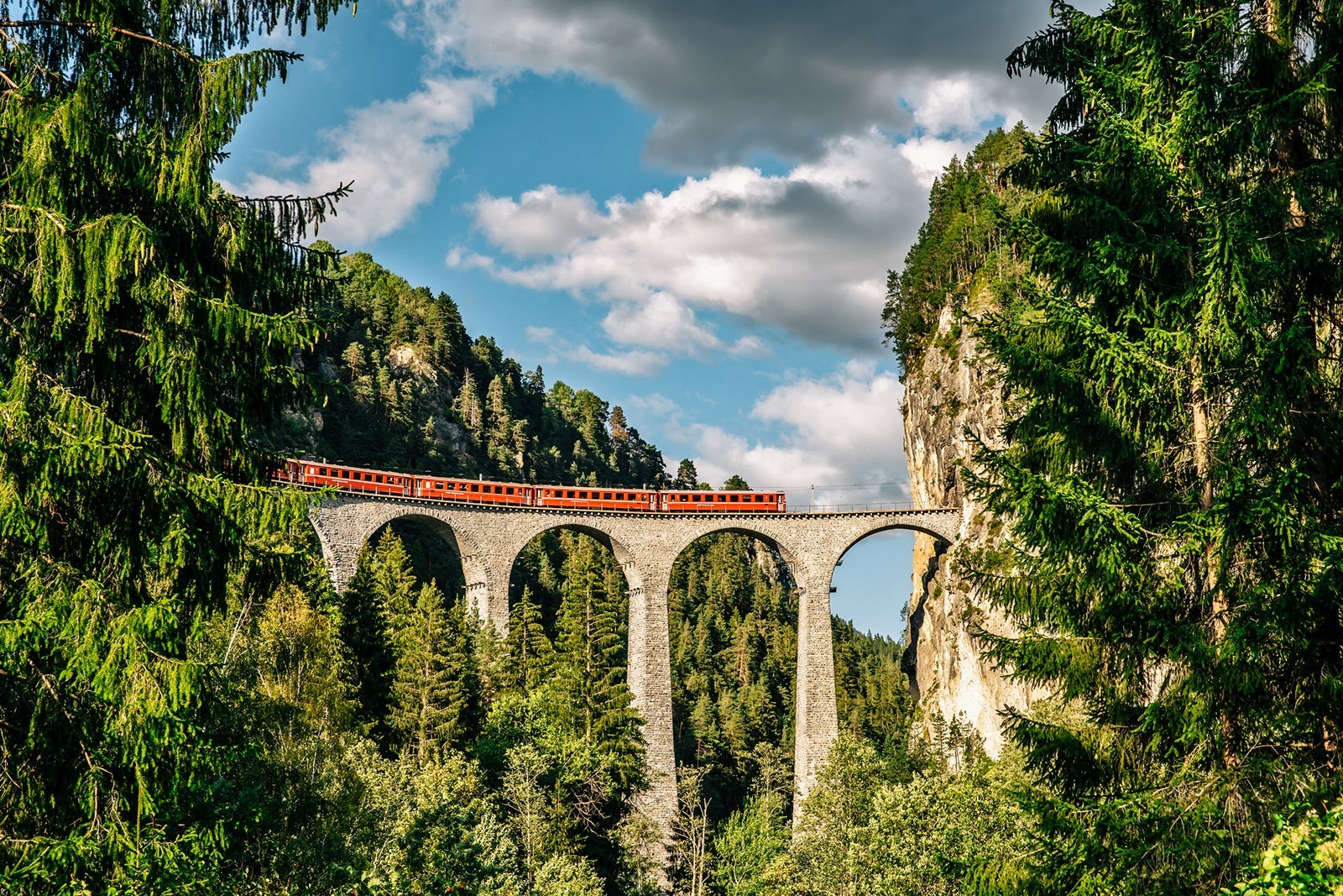 A red train crossing the high stone arches of the Landwasser Viaduct in Switzerland.